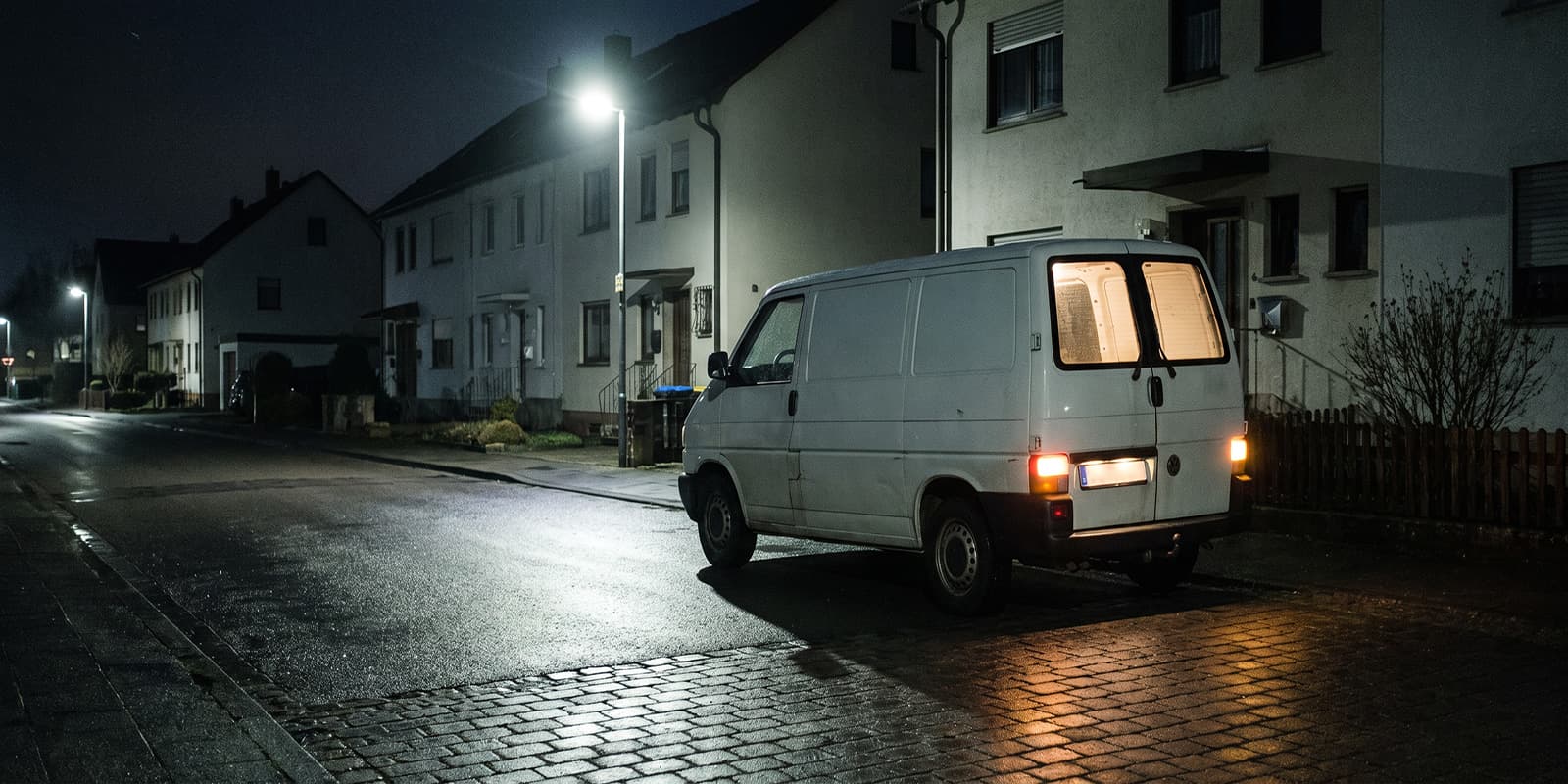 Weißer Kastenwagen nachts am Straßenrand vor einem Reihenhaus, nasse Fahrbahn, KI-generiertes Beispielbild